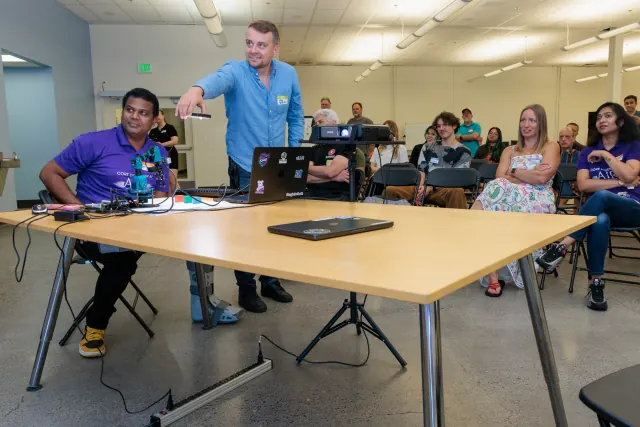 Two builders at the demo table with a small robot arm and a laptop, one pointing a remote at the screen, while the seated audience looks on smiling.