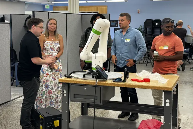 Five people wearing name tags stand talking around a small robot arm on a workbench; one holds a can of sparkling water.