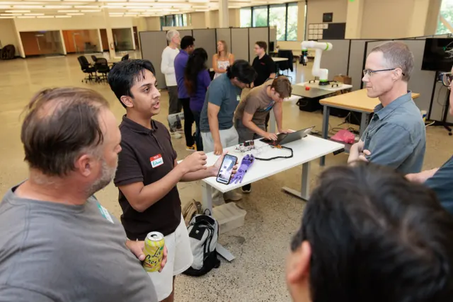 A builder holds up a phone while explaining a 3D-printed robotic hand to three listeners; behind them, others lean over the hand and its wiring on a table.